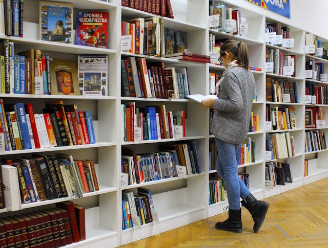 Slika prikazuje dekle, ki stoji v knjižnici pred knjižnimi policami in bere knjigo